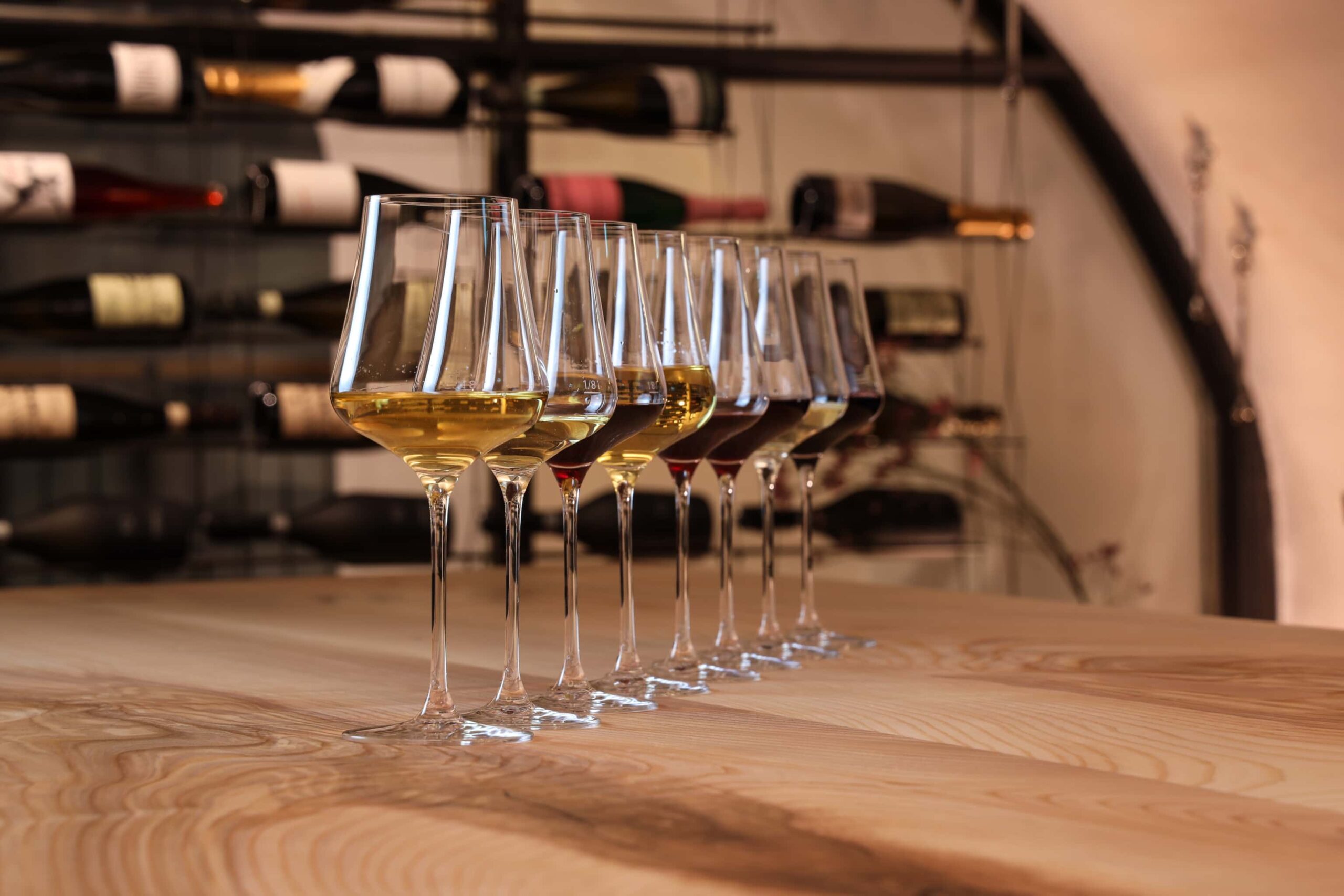 Bild-Header: Close up von acht gefüllten Weingläsern, die diagonal aufgereiht am Tisch stehen. Im Hintergrund sind verschwommen Schaumweine zu erkennen.