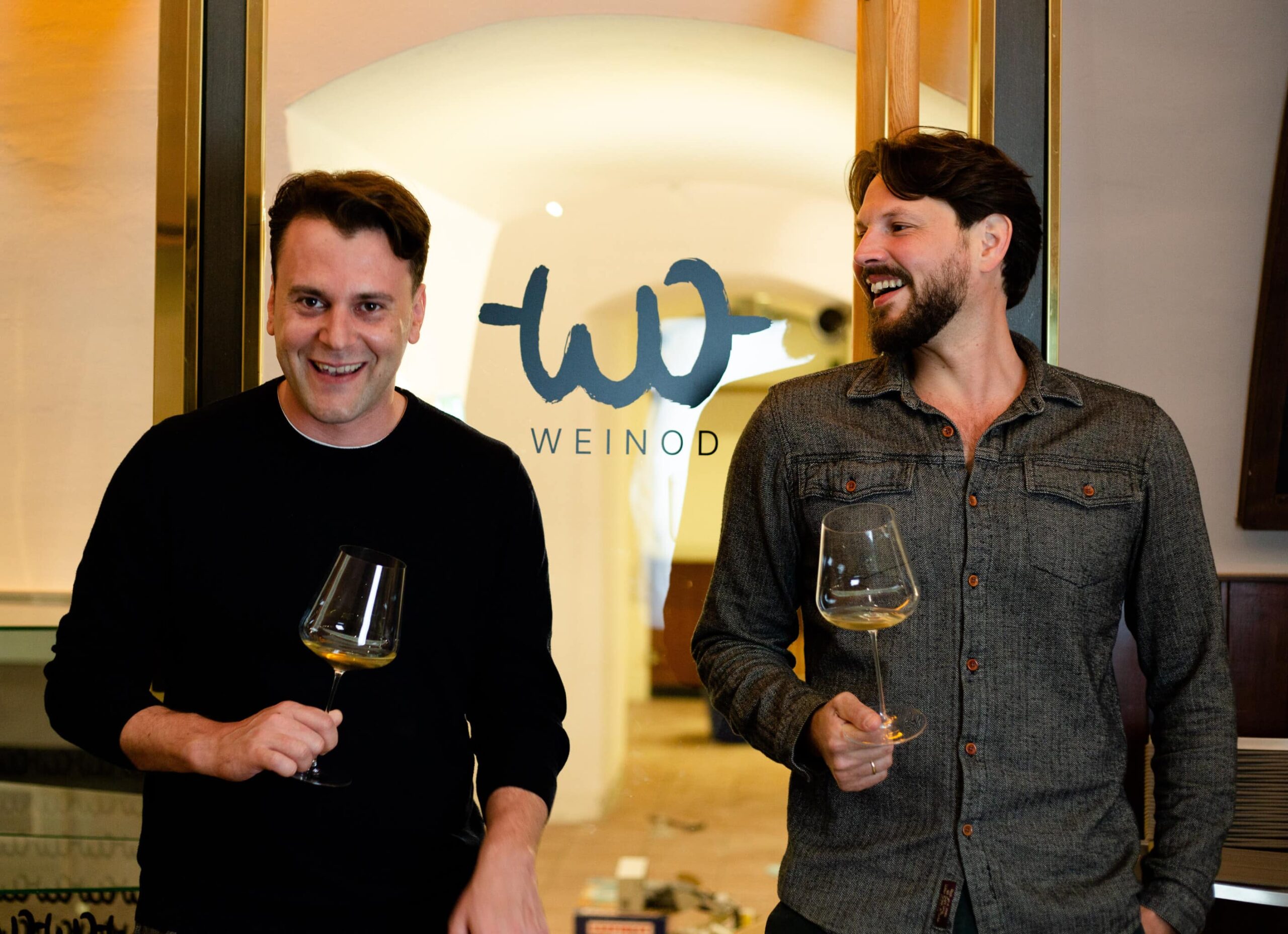 Header-Bild: Mathias und Stefan stehen mit einem Weinglas in der Hand vor dem WEINOD Eingang und freuen sich.